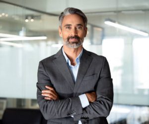 Handsome hispanic senior business man with crossed arms smiling at camera. Indian or latin confident mature good looking middle age leader male businessman on blur office background with copy space.